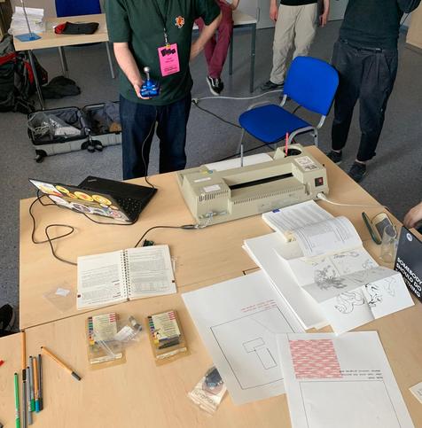 Photo of a desk with a plotter and laptop with misc. printout papers. A person is holding a joystick.