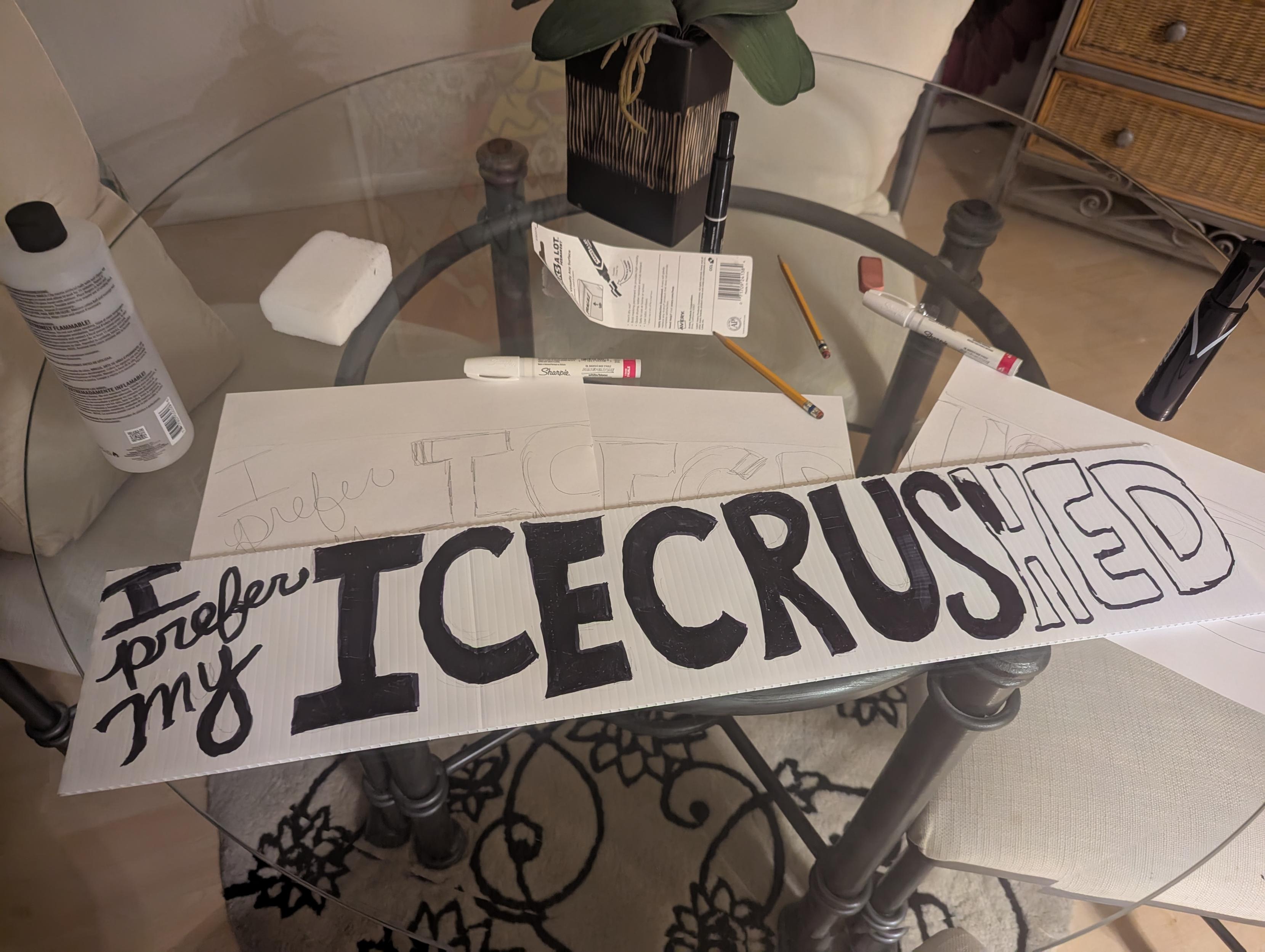 Photo of a kitchen table with craft supplies and a protest sign that says "I prefer my ICE CRUSHED"  partially colored in. 