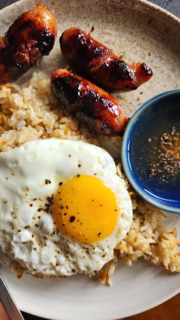 On a plate: a serving of garlic fried rice topped with fried egg, three pieces of sweet sausages, and some vinegar dipping sauce