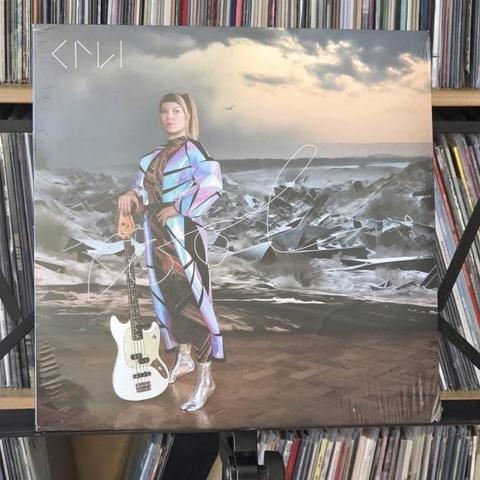 Ein Schallplattencover mit einer Person, die in einer surrealen Landschaft mit einem abstrakten, wirbelnden Hintergrund steht. Die Person ist in einem bunten, reflektierenden Outfit gekleidet und hält eine weiße Bassgitarre. Die Szene enthält Elemente der Natur und einen bewölkten Himmel.