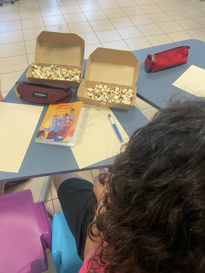 Table de classe avec boîte remplie de popcorn, feuille et stylo, le roman « bobine et popcorn » et cheveux d’un enfant