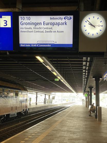 An airport train station display shows the next train to Groningen Europapark at 10:10, with connections listed. A clock indicates the current time. In the background, a train is partially visible on the platform, and a person sits on a bench