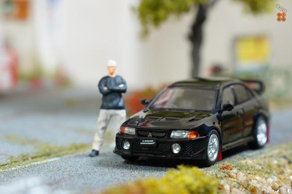 A Mitsubishi Lancer GSR Evolution V 1998 with Mud Flap, Pyrenees Black by Hobby Japan in 1:64 scale with 1:64 figure on AREA 164