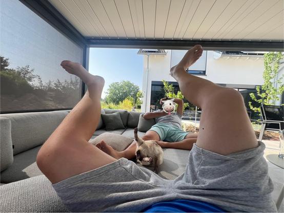 A siamese cat doing her intimate licking on the outdoor couch under a pergola, with both back legs in the air, and me next to her with my legs up in the air. My anonymized partner is laying with us at the back 