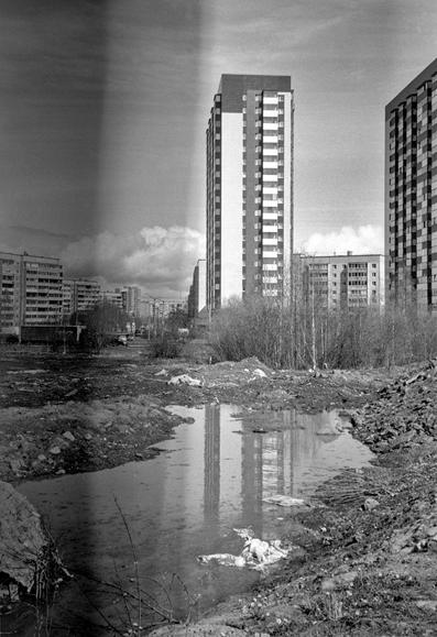 Monochrome photo shows a multi-story, new building reflected in an unsightly puddle in front of it.