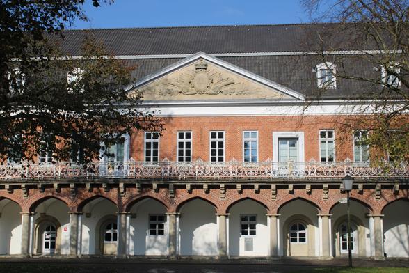 Foto vom Marstall in Aurich. Errichtet um 1588 als höfischer Pferdestall (Teil vom nebenstehenden Schloss). Das Gebäude zieht sich durch das ganze Bild, denn es ist lang und schmal. Im unteren Bereich ist ein Arkadengang, darüber befindet sich ein umlaufender "Balkon". Die Fassade besteht aus roten Ziegelsteinen. Zentral befindet sich ein Dreiecksgiebel. 