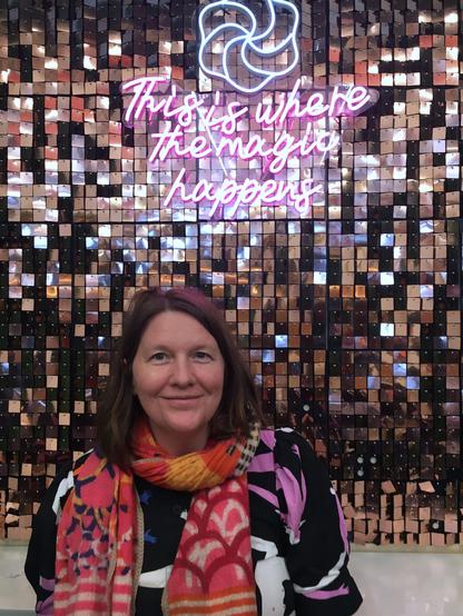 A woman poses in front of a colourful neon sign, casting a bright glow in a dark atmosphere.