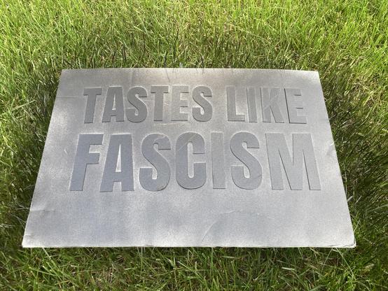 A piece of white foam core with black 3D printed letters placed on top of it which has been spray-painted.