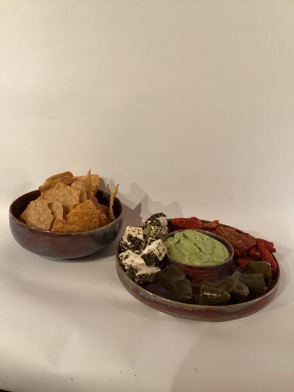 Eine Keramikschale mit Riebelchips Chili und eine Keramikschale mit Avocadodip in der Mitte und getrockneten Tomaten, Schafskäse und gefüllten Weinblättern rundherum 