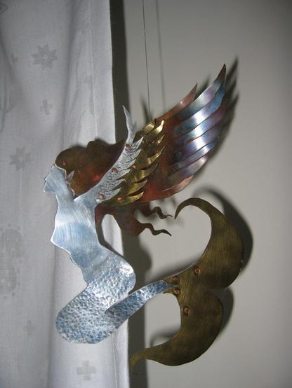 Photo of a metal sculpture of a mermaid with wings. There are different metals that are riveted and layered together. The tail is copper colored, the tail/body/face are silver, the hair is copper, and the wings from the arms are 3 different metals. The wings have feathers that are bent forward a bit to show depth.