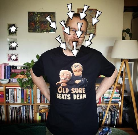 Mężczyzna w średnim wieku na tle półek z książkami. Ma na sobie czarną koszulkę że Statlerem i Waldorfem z Muppet Show i napisem: "OLD SURE BEATS DEAD!"