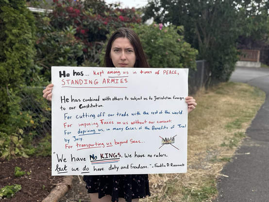 A woman holds up a protest sign quoting the Declaration of Independence grievances, “He has… kept among us in times of peace standing armies. He has combined with others to subject us to Jurisdiction foreign to our Constitution. For cutting off our trade with the rest of the world. For imposing Taxes on us without our consent. For depriving us, in many Cases of the Benefits of Trial by Jury. For transporting us beyond Seas…” Another quote follows from Franklin D. Roosevelt, “We have no kings, we have no rulers, but we do have duty and freedom.”
