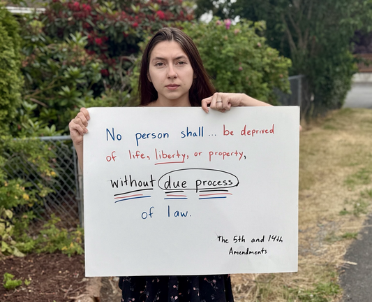 A woman holds a protest sign referencing the 5th and 14th Amendments of the United States Constitution reading “No person shall.. be deprived of life, liberty, or property without due process of law.”
