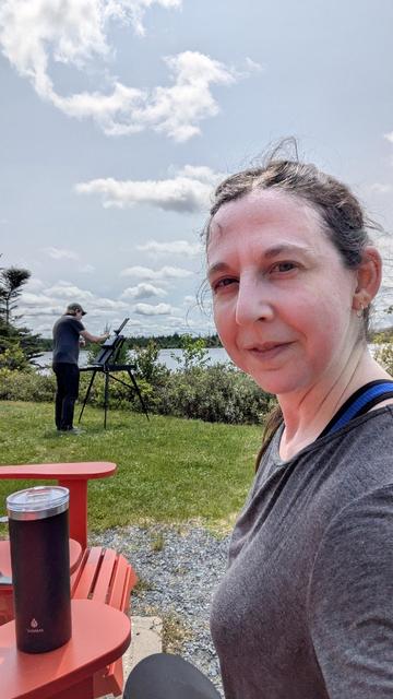 A sweaty Kyla, hair in a ponytail wearing workout clothes in front of a pond.  Her husband has an easel set up by the pond and is painting in the background.  It is a nice sunny summer day.