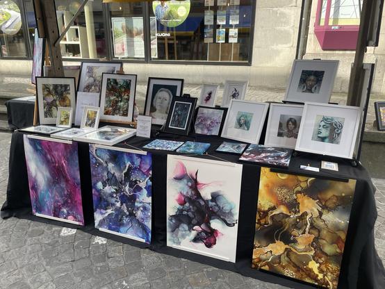 Eine Ausstellung mit verschiedenen Kunstwerken, darunter farbenfrohe abstrakte Gemälde und gerahmte Porträts. Das Kunstwerk zeigt eine Reihe von Stilen und leuchtenden Farben, die auf einem mit einem schwarzen Tuch bedeckten Tisch in einer Marktumgebung im Freien angeordnet sind.