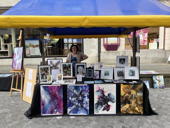 Ein Marktstand mit einer Frau, die dahinter steht und verschiedene gerahmte Kunstwerke und Drucke ausstellt. Der Stand ist von einem blau-gelben Baldachin bedeckt. Die Kunstwerke zeigen lebendige Farben und abstrakte Designs, mit einer Vielzahl von Stilen, die auf einer schwarzen Decke präsentiert werden.