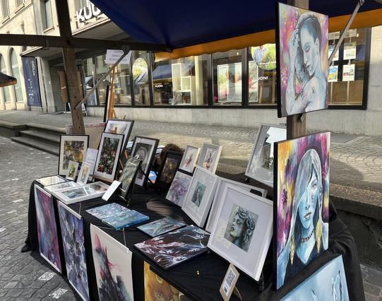 Ein Straßenkunstmarktstand zeigt verschiedene gerahmte Kunstwerke, darunter farbenfrohe Porträts und abstrakte Gemälde, die unter einem blauen Baldachin stehen. Der Hintergrund zeigt Gebäude und Sitzgelegenheiten im Freien.