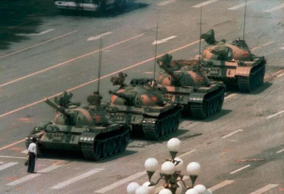 A lone man in a white shirt and dark trousers stands in front of a line of four military tanks on a wide, empty boulevard, blocking their path. The tanks are in a straight line, facing the man, and appear to be part of a military convoy. The setting is urban, with street markings and a decorative lamp post visible in the foreground. The image is a historic moment symbolizing protest and resistance.