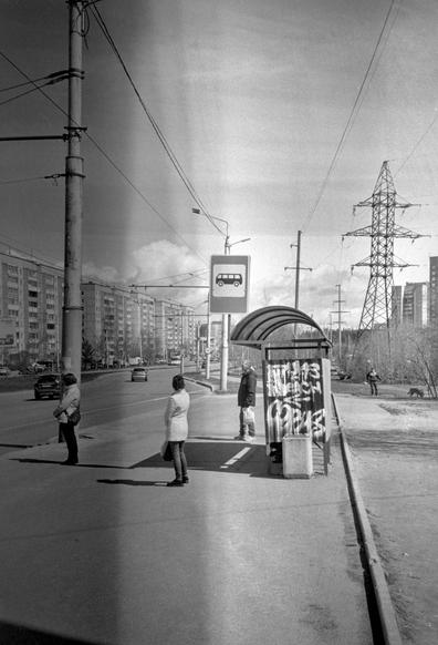 A monochrome photo shows people standing at a bus stop. Contrasting shadows fall from the people and from the bus stop.  The side of the bus stop is painted with graffiti. Behind the bus stop is a panorama