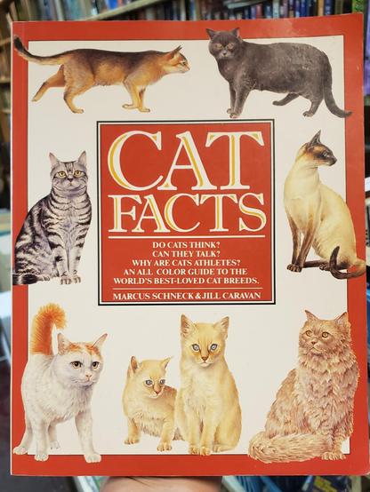 A photo of the front of a large softcover book with a red border. The book is "Cat Facts" by Marcus Schneck and Jill Caravan. A red box in the middle of the cover had a thin black border and contains title and author info with white text that reads, "Do cats think? Can they talk? Why are cats athletes? An all-color guide to the world's best loved cat breeds. The space between the red border and the red box is white with illustrations of seven different cat breeds.