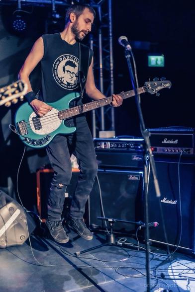 A musician playing an electric bass guitar while jumping on stage, wearing a sleeveless black shirt and dark pants. The setting features amplifiers in the background and stage lighting, creating a vibrant concert atmosphere.
