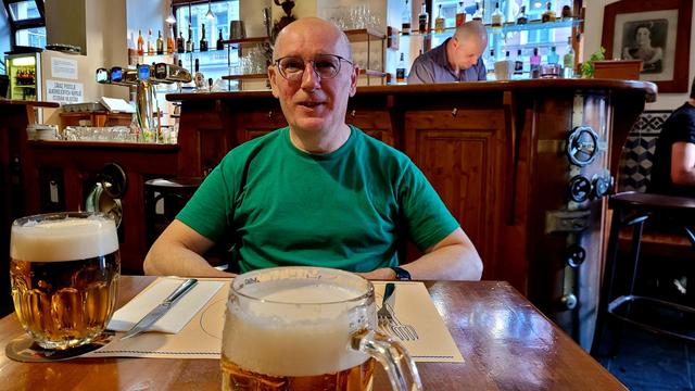 Mann im grünen T-Shirt sitzt an einem Restaueanttisch, auf dem 2 Bierkrüge stehen. Die Theke ist mit Industriemaschinenteilen dekoriert.