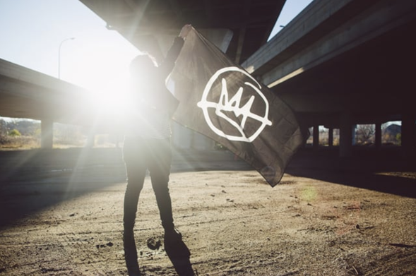 person under a bridge, obscured by the sun, waving a black No Kings flag