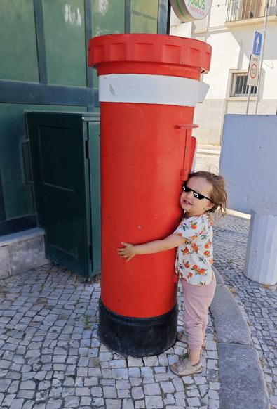A young child wearing sunglasses hugs a large red cylindrical mailbox on a cobblestone sidewalk, smiling happily.