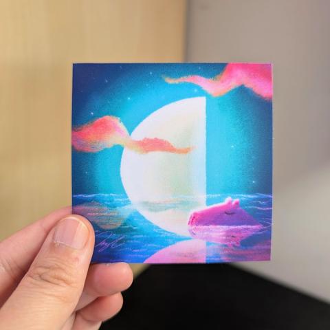 Photo of a left hand holding a square sticker. The sticker is a painting of a capybara resting in water with a half moon in the background.