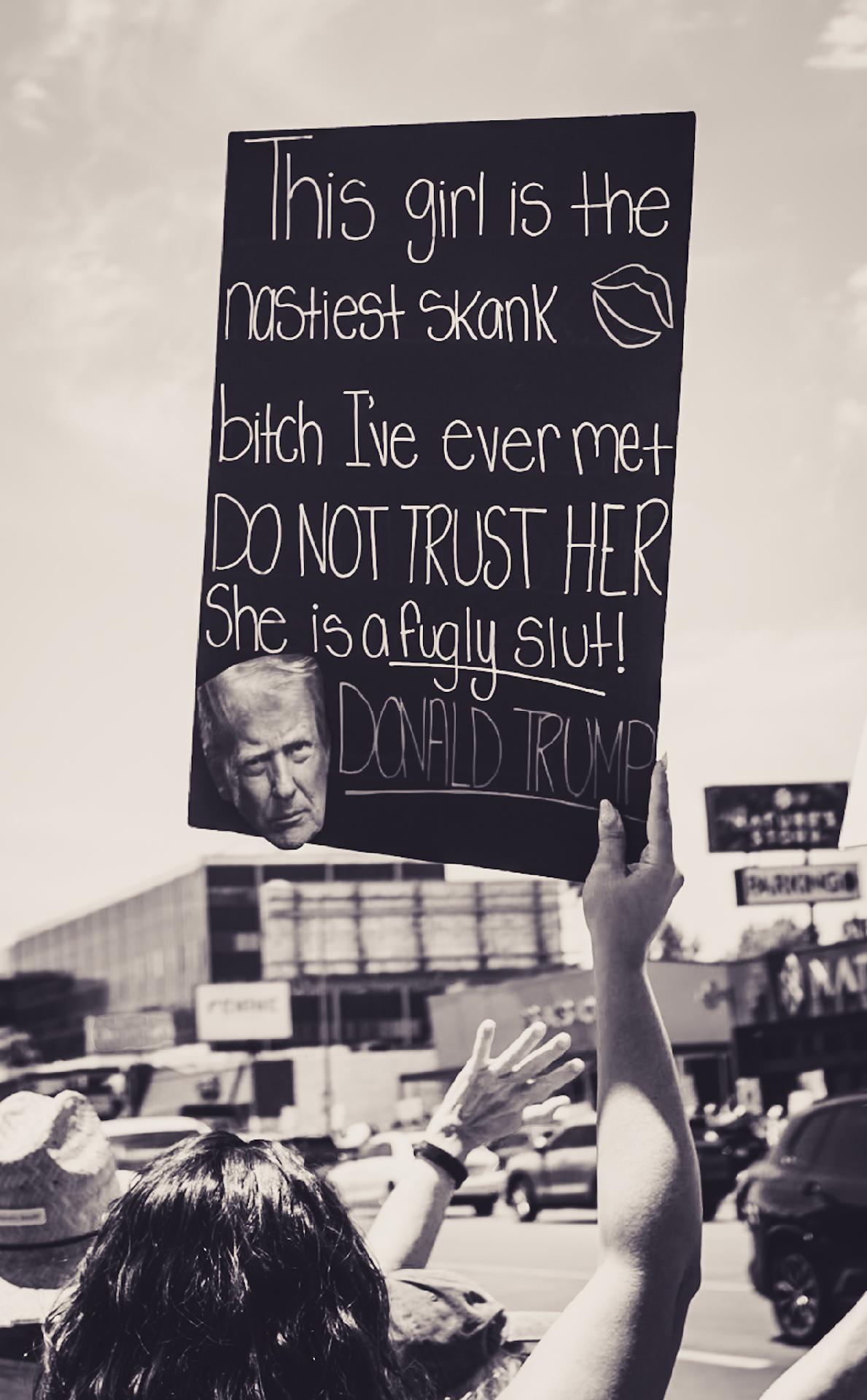 A protester holding a sign with Trump's face that reads: This girl is the nastiest Skank bitch I've ever met  DO NOT TRUST HER  She is a fugly Slut