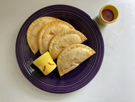 A dark purple plate holds four empanadas. There are two yellow shot glasses filled with hot sauce. A decal on the yellow shot glasses is of a chili pepper.