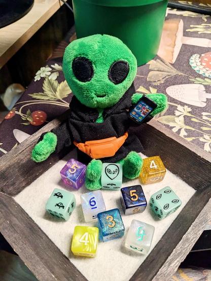 A small green alien doll, wearing a black hoodie and an orange fanny pack, holding a tiny cell phone in her left hand. She is sitting in a square, wooden dice tray surrounded by several different six-sided dice in various styles.