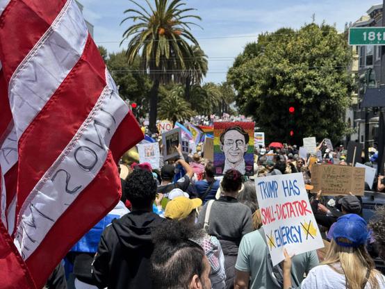 A large crowd of folks at the No Kings protest in San Francisco. Protestors are waving American flags and protest signs. One sign says “Trump has small dictator energy” another has a painting of Harvey Milk and a rainbow background. 