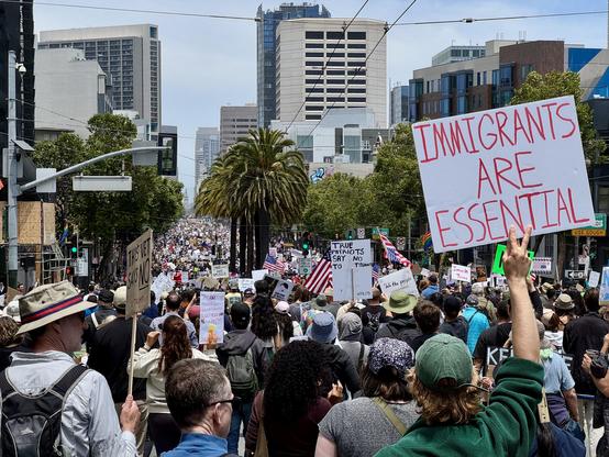 Another photo of the large crowd this one is taken from on top of a hill on Market St, so you can see thousands of attendees going for blocks and blocks. A sign in the foreground says “Immigrants are essential”. 