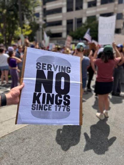 Protest sign saying “SERVING NO KINGS SINCE 1776” with the words between two buns, thanks to buns.life
