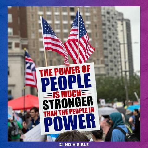 A protester holds a sign reading "The power of people is much stronger than the people in power" with two American flags in the background, at an Indivisible rally.