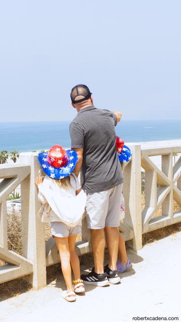 A man and his two young daughters wearing star spangled cowboy hats look out over the ocean 