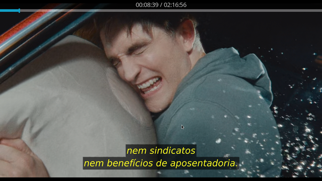 A imagem mostra um homem em um carro, com a cabeça apoiada num airbag, parecendo estar em dor ou angústia. Ele está usando um moletom cinza e tem uma expressão de sofrimento no rosto. A janela do carro está aberta, e há cacos de vidros visíveis ao redor, sugerindo que ele está no carro que acabou de sofrer um acidente. No canto superior direito, há uma barra de progresso de vídeo com o tempo atualizado em "00:08:39 / 02:16:56". Na parte inferior da imagem, há uma legenda em português que diz: "nem sindicatos nem benefícios de aposentadoria."