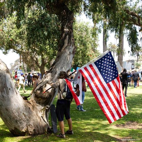 A man with a large American flag and a California flag getting ready to ride his skateboard 
