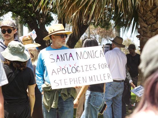 A man holds a sign that says “Santa Monica apologizes for Stephen Miller”