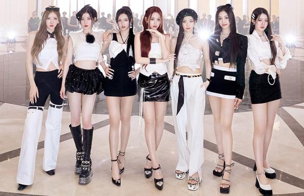 The 7 members of BABYMONSTER pose for a promotional photo in the clothes and set of FOREVER's music video. They're all dressed and black and white, some of them in pants and others in short skirts or dresses. Behind her, a crowd is gathered and looking at them with admiration and/or curiosity. The background is a light beige color and lit extremely bright, to bring the attention to the girls, but also giving it a sort of light gold appearance. There's a very high end aspect to this picture, enhanced by the fact that the music video includes scenes on a fashion runway.