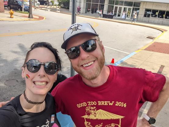 Selfie photo of myself and my partner. I'm wearing a bisexual color skeleton holding coffee black T-shirt. My partner is wearing a red Good Zoo Brew 2014 T-shirt. 