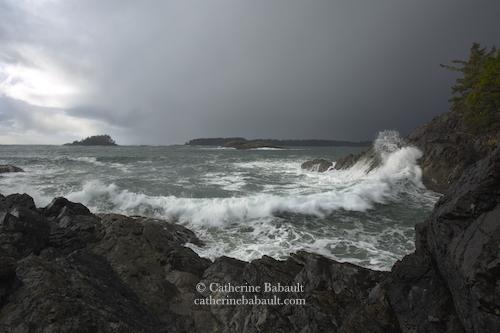 Big waves crashing on a rocky shoreline. The sea is agitated and has a green tint, there are islets on the horizon. The sky is dark.