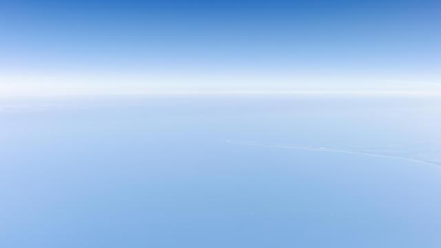 View from an airplane. Very bright view of the North Sea and blue sky, with a thin cloud layer roughly on the horizon. A long piece of land in Northern Denmark sticking out into the sea.