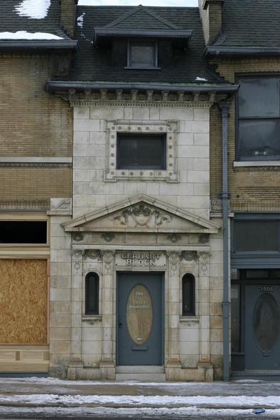 A color photo of a row of stone and brick buildings. Centered in the frame is a white stone building with a somewhat ornate facade and a black roof. Above the door, the stone is carved to read "GERHART BLOCK". The street and sidewalk in front of the building is snowy.