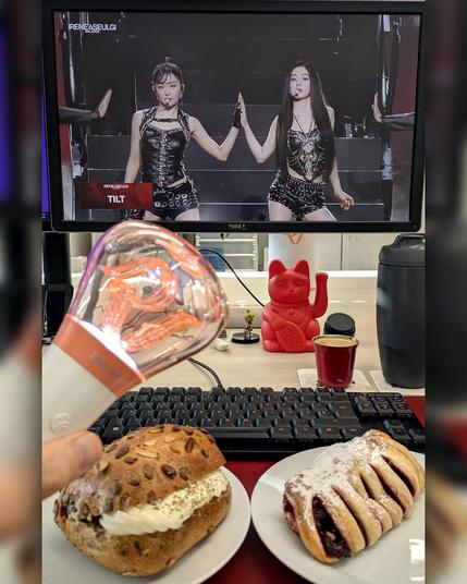 picture of an office desk, the monitor is showing a concert video and there is breakfast in front of the keyboard. a hand is holding a kpop cheering lightstick.