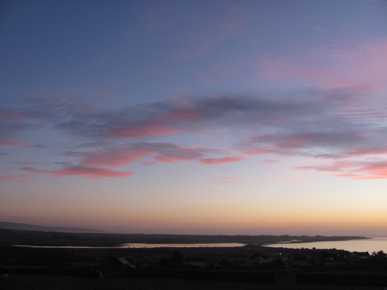 Late sunset - the sky is darkening in tones of deep yellow- orange at the horizon, to deep violet at the top, with clouds in pink and indigo. The coastline below is dark