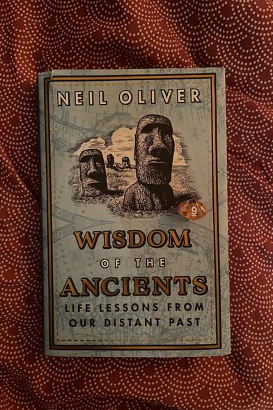 A book on a red and white patterned bedspread. A yellow D10 sits on top of the book showing the number 9. 

The book cover shows the Moai, NEIL OLIVER
WISDOM OF THE ANCIENTS; LIFE LESSONS FROM OUR DISTANT PAST