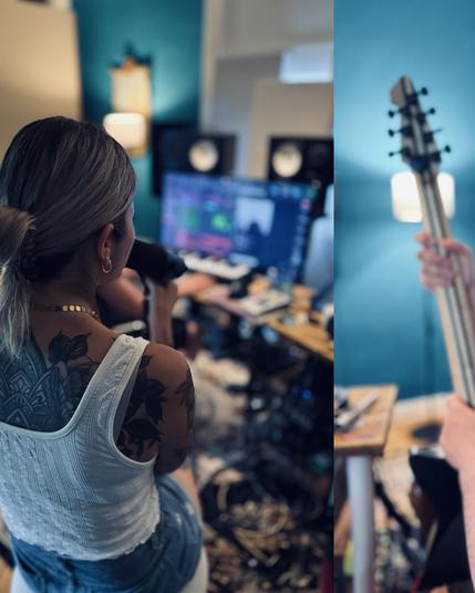 Das Bild zeigt Kim, Sängerin der Band DARKK mit silbergrauem Haar, das zu einem Pferdeschwanz gebunden ist, die von hinten in einem Musikstudio sitzt. Sie trägt ein weißes Tanktop und hat eine große, detaillierte Tätowierung auf dem Rücken und Arm. Sie hält ein Mikrofon in der Hand und singt in die Kamera. Im Hintergrund ist ein Computer mit einem Musikproduktionstool sichtbar, sowie Lautsprecher und andere Musikproduktionsequipment. Auf der rechten Seite des Bildes ist ein Bassgitarrenkopf zu …