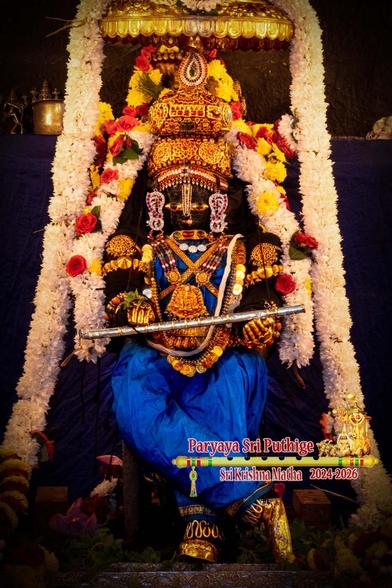 Idol of Hindu deity Krishna in blue standing cross-legged (right leg over left); wearing gold shoes, gold crown, and several gold ornaments; holding a silver flute (held almost horizontal) in the palm of his hands; a small gold umbrella overhead.

A thick string of white flowers from Krishna's left waist going up over his crown and falling down the other side to his right waist. A similar thick string of white flowers goes from the floor and over the umbrella, flowing on either side of Krishna.